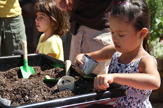 a little girl scoops soil into a paper planter, with other people in the background